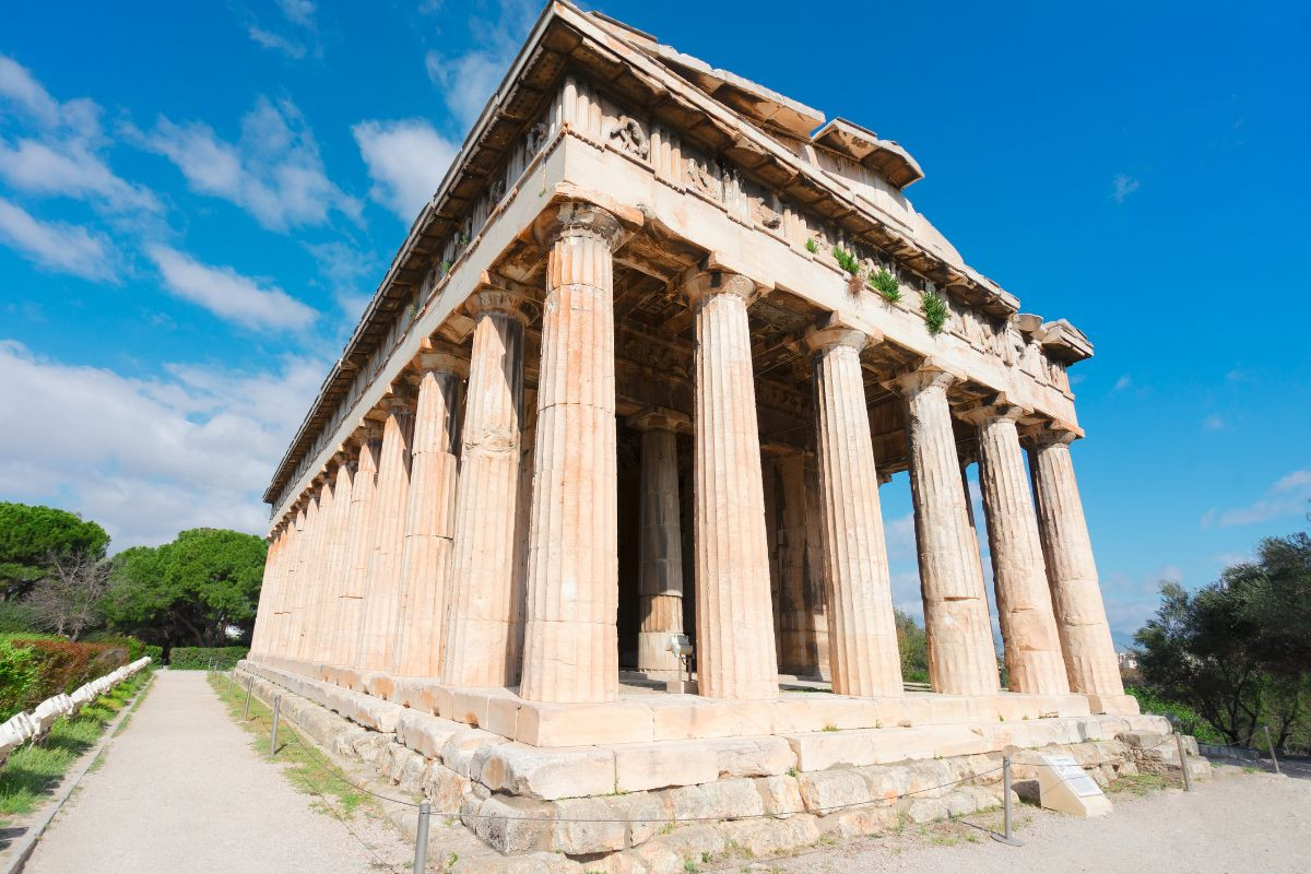 The image shows the Temple of Hephaestus in the ancient Agora of Athens. The temple, built in classical Greek architectural style, features well-preserved Doric columns and is set against a blue sky with some clouds.